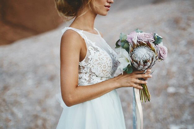 Belle fille blonde modèle avec modélisation de coiffure de mariage dans une robe en dentelle blanche à la mode avec un bouquet de fleurs exotiques dans ses mains