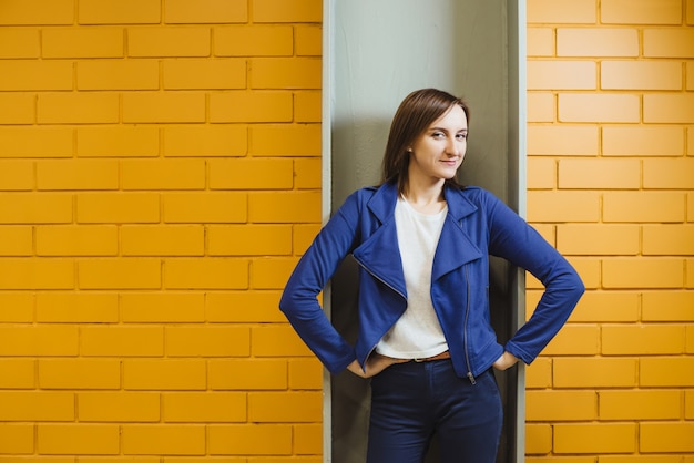 Belle fille blonde moderne gaie dans une entreprise pose contre un mur de brique jaune.