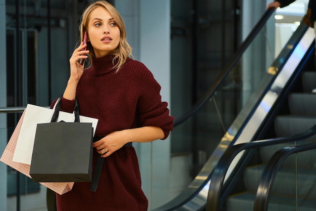 Photo belle fille blonde en pull tricoté parlant en toute confiance sur un téléphone portable dans un centre commercial moderne