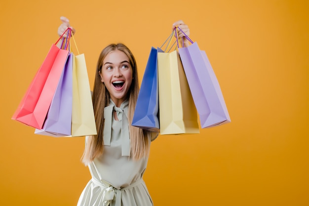 Belle fille blonde souriante heureuse avec des sacs à provisions colorés isolé sur jaune