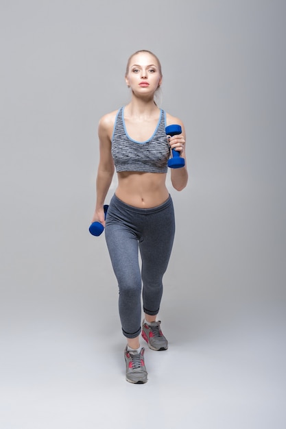 belle fille blonde en tenue de sport fait des exercices avec des haltères sur fond gris