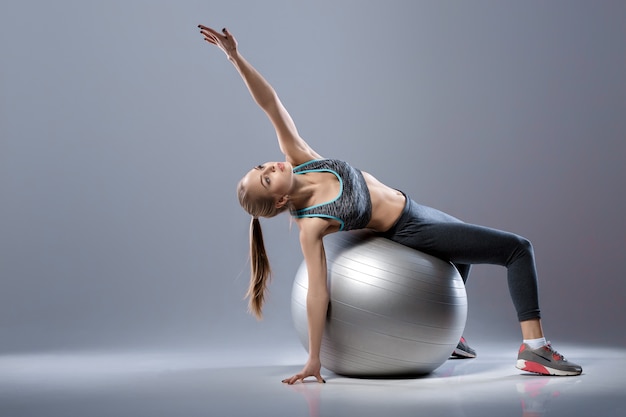 belle fille blonde en vêtements de sport assis sur un ballon de fitness sur fond sombre