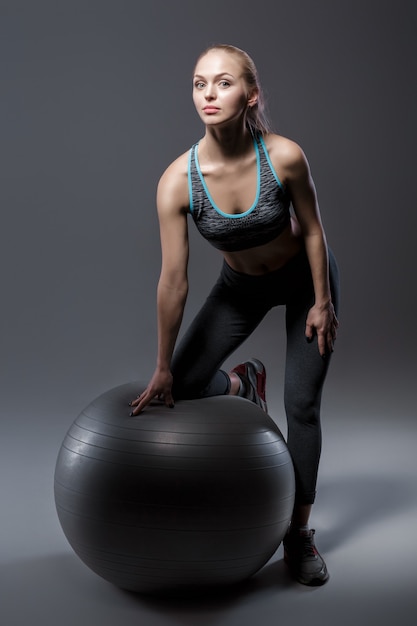 belle fille blonde en vêtements de sport assis sur un ballon de fitness sur fond sombre