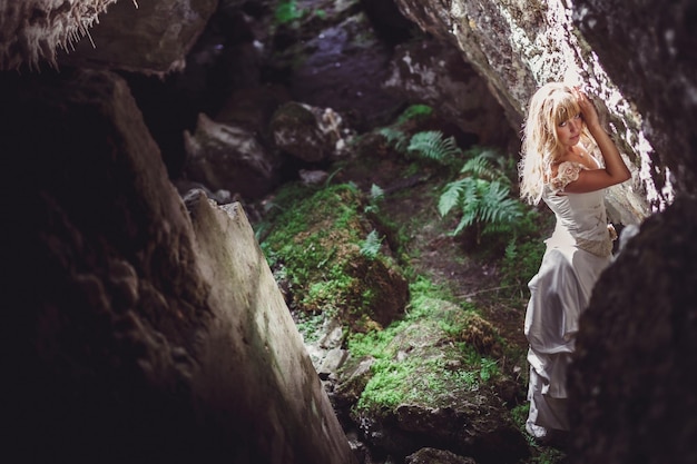 Belle fille blonde vêtue d'une robe blanche avec un corset ou un pantalon en cuir dans une grotte sombre et profonde