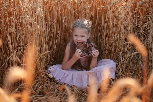 une belle fille blonde vêtue d'une robe en lin rose est assise dans un champ de blé et mange du pain