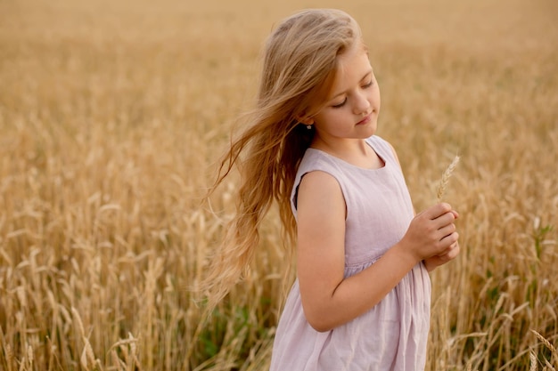 belle fille blonde vêtue d'une robe en lin rose se tient dans un champ de blé et la touche avec ses mains