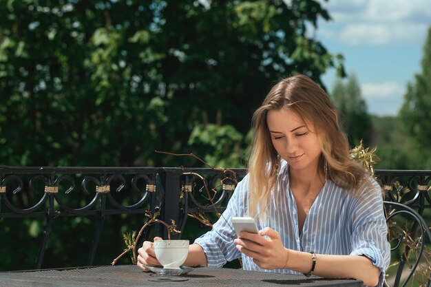 Belle fille boit du café à une table sur le balcon à la maison et regarde à travers son téléphone