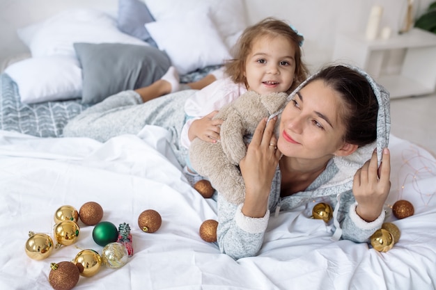 Une belle fille bouclée de 2 ans avec sa jeune mère s'allonger sur le lit parmi les guirlandes et les décorations et rire