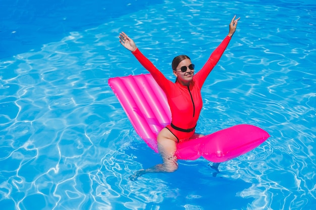 Belle fille bronzée en maillot de bain rose et lunettes au bord de la piscine Tient un matelas pneumatique rose lunettes de soleil peau bronzée humide Station de vacances plage Journée ensoleillée