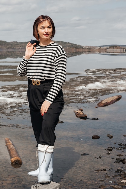 Belle fille brune au bord de la rivière. Une fille vêtue d'un gilet rayé et de bottes en caoutchouc tient une veste à la main. Loisirs et tourisme.