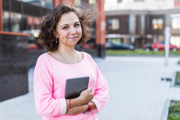 Une belle fille brune aux vêtements à la mode est debout et tient une tablette à l'extérieur en été.