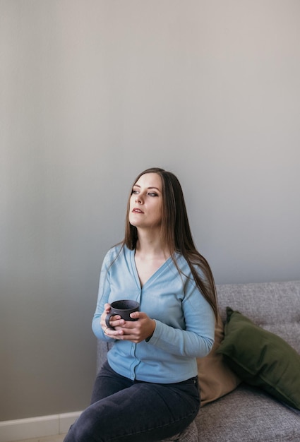 une belle fille brune boit du thé dans une tasse sur le canapé de la chambre et regarde par la fenêtre