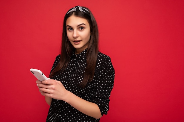 Belle fille brune debout isolée sur un mur rouge portant des vêtements noirs élégants et décontractés