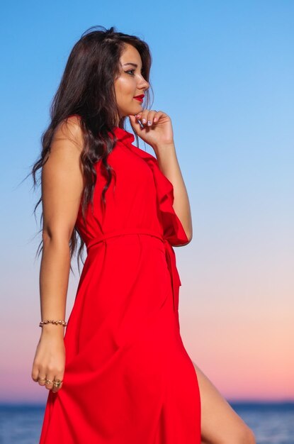 Belle fille brune en robe rouge posant au bord de la mer au lever ou au coucher du soleil