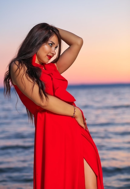 Belle fille brune en robe rouge posant au bord de la mer au lever ou au coucher du soleil