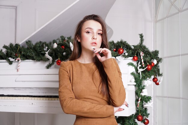 Belle fille brune sensuelle aux longs cheveux raides près d'un piano blanc avec décoration de Noël