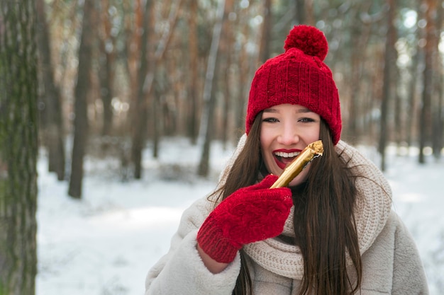 Belle fille brune souriante à l'extérieur en bonnet rouge tricoté et des mitaines avec des bonbons dans ses mains. Femme dans la forêt d'hiver
