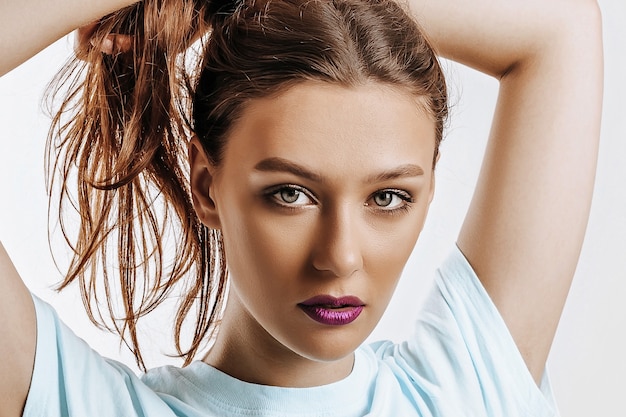 Une belle fille brune tient ses cheveux et fait sa propre coiffure. Jeune mannequin aux cheveux longs, maquillage mode aux lèvres violettes sur fond gris. La femme regarde sérieusement la caméra.