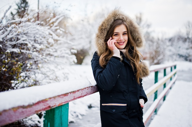Belle fille brune en vêtements chauds d'hiver. Modèle sur la veste d'hiver contre le lac gelé au parc parle au téléphone mobile.
