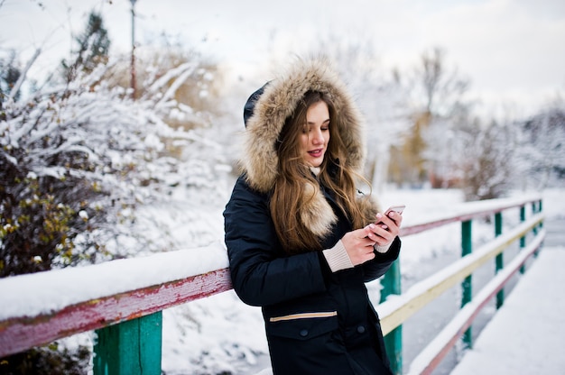 Belle fille brune en vêtements chauds d'hiver. Modèle sur veste d'hiver contre lac gelé au parc parler sur téléphone mobile.