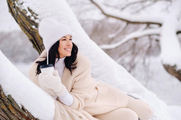 Une belle fille avec un cardigan beige et un chapeau blanc appréciant de boire du thé dans une forêt d'hiver enneigée près d'un lac