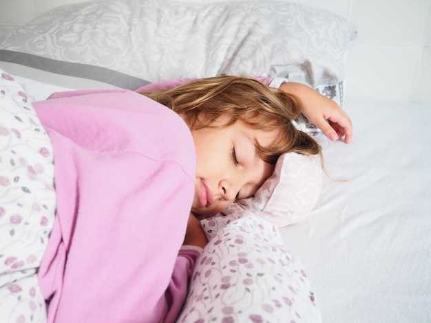 Une belle fille caucasienne de 8 ans aux cheveux blonds vêtue d'un pyjama rose dort sur un lit avec une couverture moelleuse serrant un oreiller La douce lumière du soleil du matin traverse la fenêtre