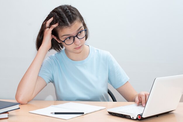 Belle fille caucasienne est assise à un bureau avec des accessoires scolaires et a l'air surpris d'un ordinateur portable