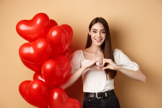 Belle fille caucasienne exprime l'amour et la tendresse, montrant le geste du cœur et souriant, debout près des ballons de la Saint-Valentin, fond beige.