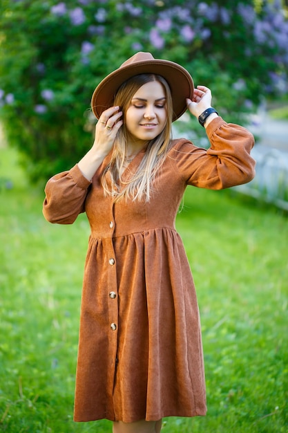 Photo belle fille avec un chapeau brun debout près d'un buisson de lilas. belle fille au chapeau près d'un arbre en fleurs.