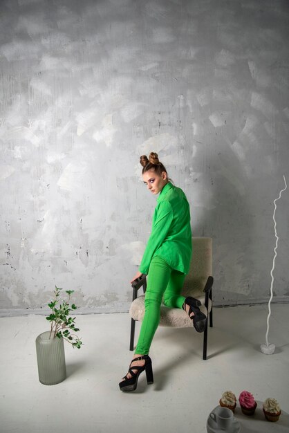 Belle fille en chemise verte et pantalon sur chaise sur fond de mur gris Femme élégante photographiée en studio Cadre vertical