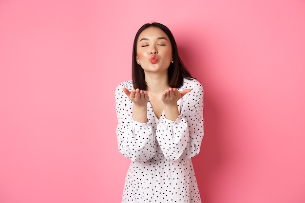 Belle fille chinoise en robe soufflant des baisers d'air, envoyant mwah à la caméra, debout sur le rose.