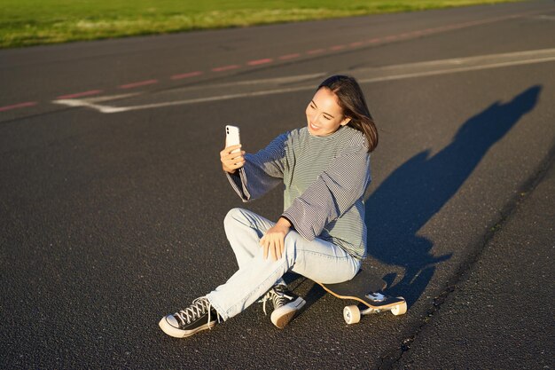 Une belle fille coréenne prend un selfie sur un smartphone prend une photo avec son skateboard profite d'une journée ensoleillée