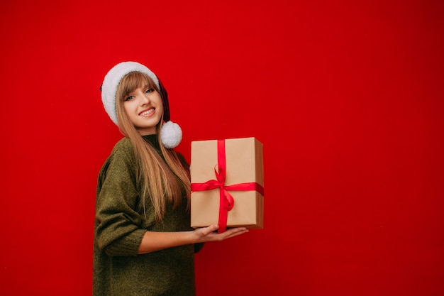 Une belle fille dans un bonnet de Noel tient un cadeau du Nouvel An dans ses mains sur un fond rouge Noël