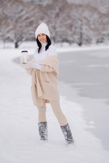 Une belle fille dans un cardigan beige et un chapeau blanc avec un verre de thé profite d'un talus enneigé au bord du lac