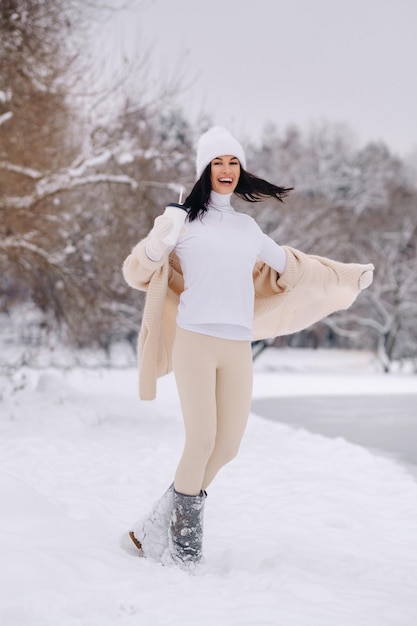 Une belle fille dans un cardigan beige et un chapeau blanc avec un verre de thé profite d'un talus enneigé au bord du lac