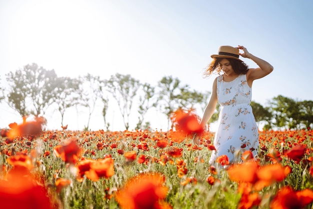 Belle fille dans le champ de pavot au coucher du soleil dans une robe blanche et un chapeau