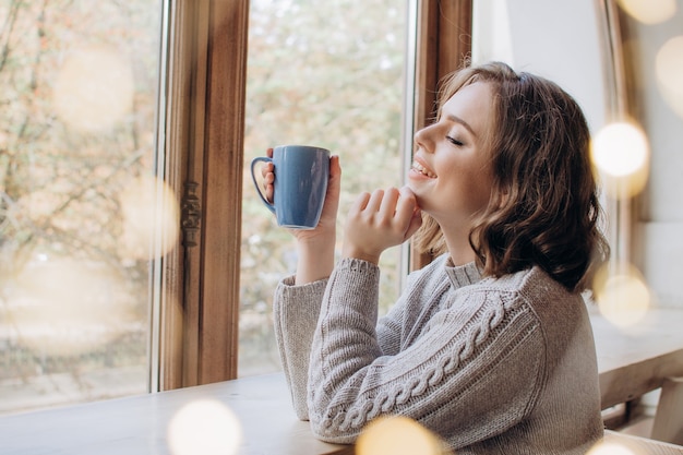 Une belle fille dans un chandail boit du thé ou du café dans le contexte d'une fenêtre et de lumières