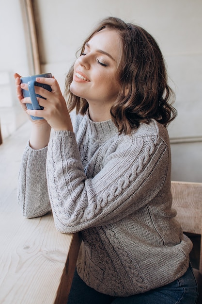 Une belle fille dans un chandail boit du thé ou du café sur le fond de la fenêtre