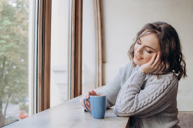 Une belle fille dans un chandail boit du thé ou du café sur le fond de la fenêtre