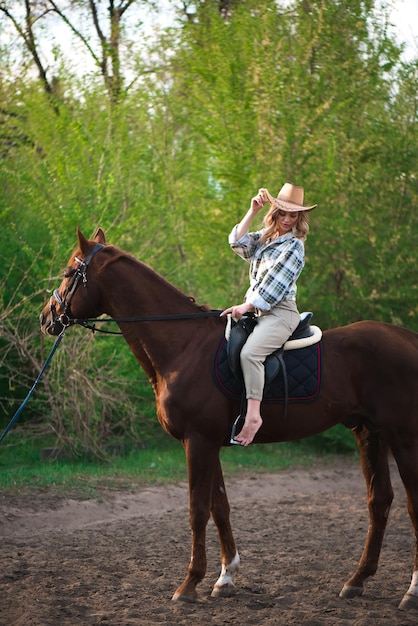 Belle fille dans un chapeau montant un cheval dans la campagne