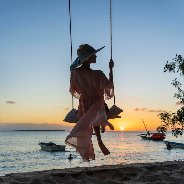 Belle fille dans un chapeau de paille et un paréo se balançant sur une balançoire sur la plage pendant le coucher du soleil de l'île de Zanzibar Tanzanie Afrique de l'Est Concept de voyage et de vacances