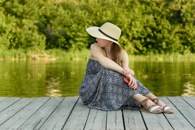 Belle fille dans un chapeau et une robe est assise sur une jetée au bord de la rivière