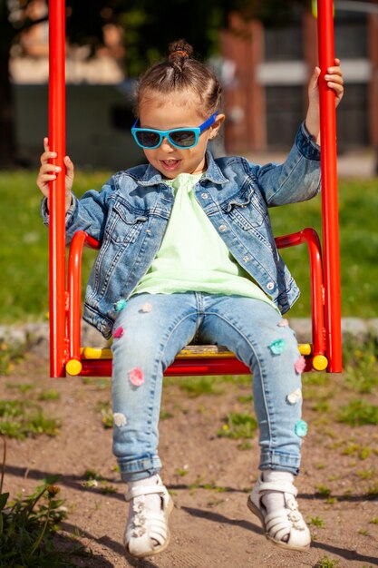 Belle fille dans un costume en jean et des lunettes de soleil se balançant sur une balançoire