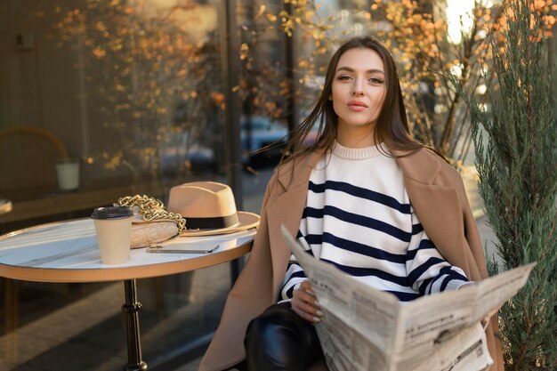 Belle fille dans un manteau lit un journal et sourit tout en se relaxant dans un café en plein air