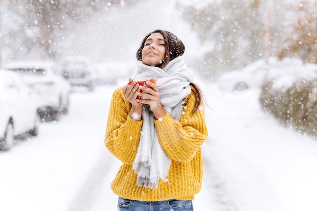 Belle fille dans un pull jaune et une écharpe blanche debout avec une tasse rouge dans une rue enneigée