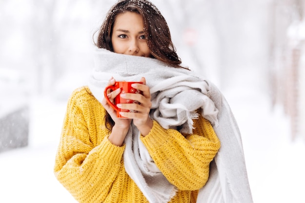 Belle fille dans un pull jaune et une écharpe blanche debout avec une tasse rouge dans une rue enneigée