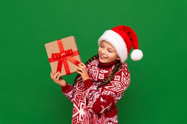 Une belle fille dans un pull de Noël et un chapeau de Père Noël tient un cadeau du Nouvel An dans ses mains Un enfant avec des nattes et un pull d'hiver chaud avec un cadeau dans ses mains