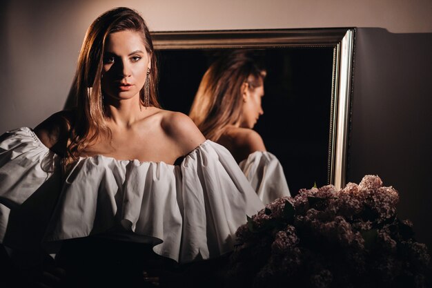 Belle fille dans le reflet du miroir à la maison. Fille avant les vacances près du miroir de la maison. Une fille en robe blanche aux cheveux longs va à une fête à la maison