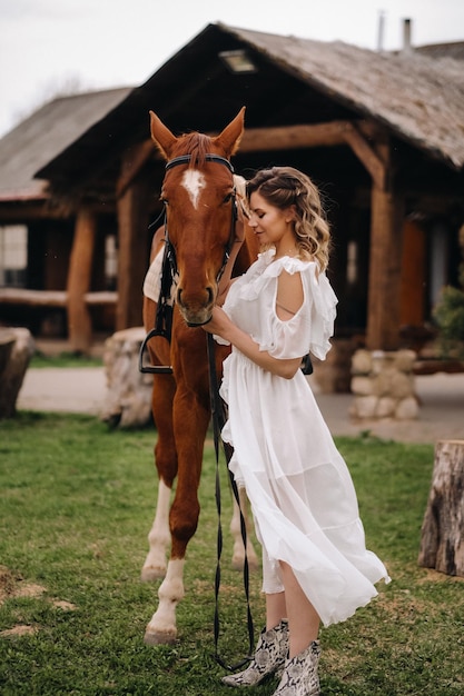 Belle fille dans une robe d'été blanche à côté d'un cheval sur un vieux ranch