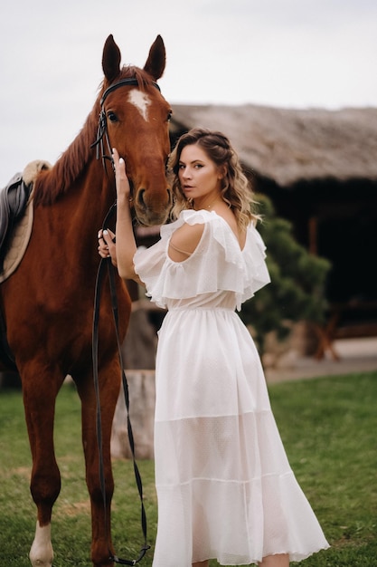 Belle fille dans une robe d'été blanche à côté d'un cheval sur un vieux ranch